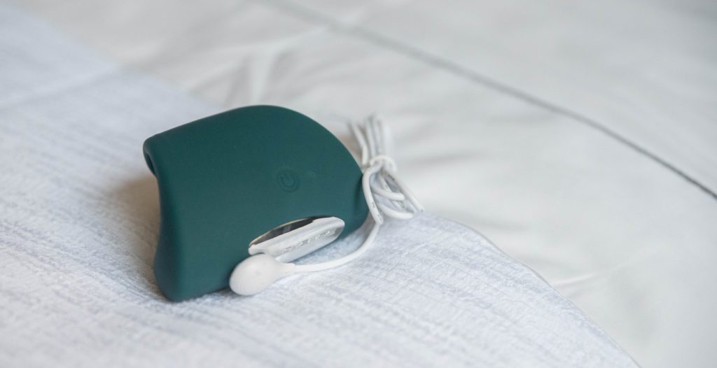 The Lovense Gush sits out on the white bedding of a bed. The magnetic charging cable is connected to the Gush to show how the charging cable connects to the toy. The opposite end of the charging cable is a USB connector. For my Lovense Gush review.