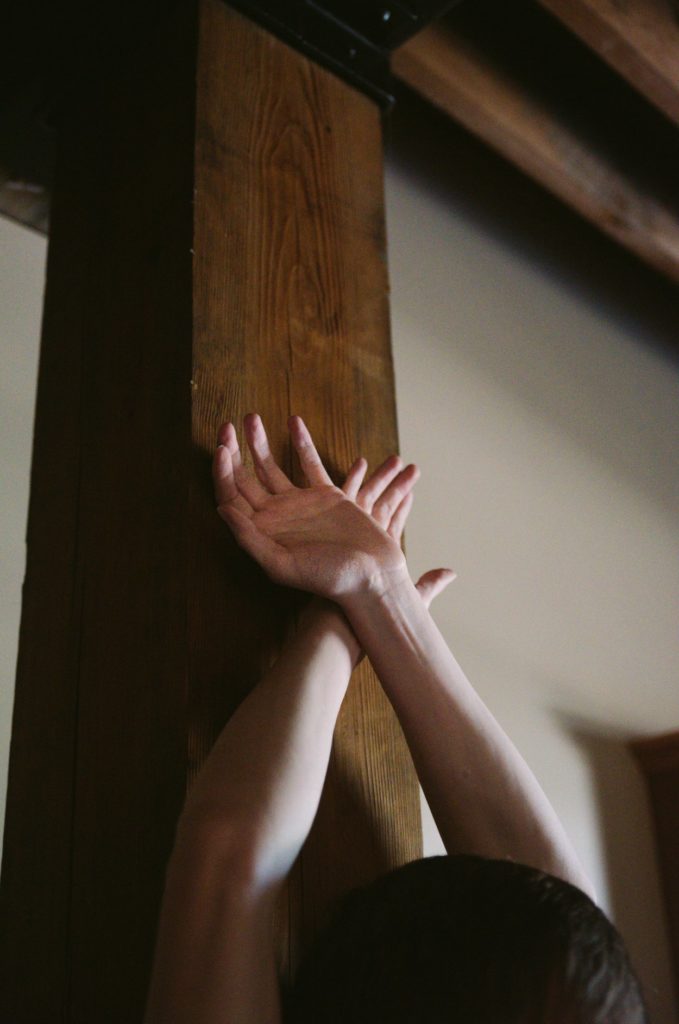Two hands gripping a large, wooden pole. There are no cuffs or any other kink items, but the person leaves their hands up in this odd position against the wooden pole. The image is shadowed and moody. Image for Mental Bondage in BDSM. 