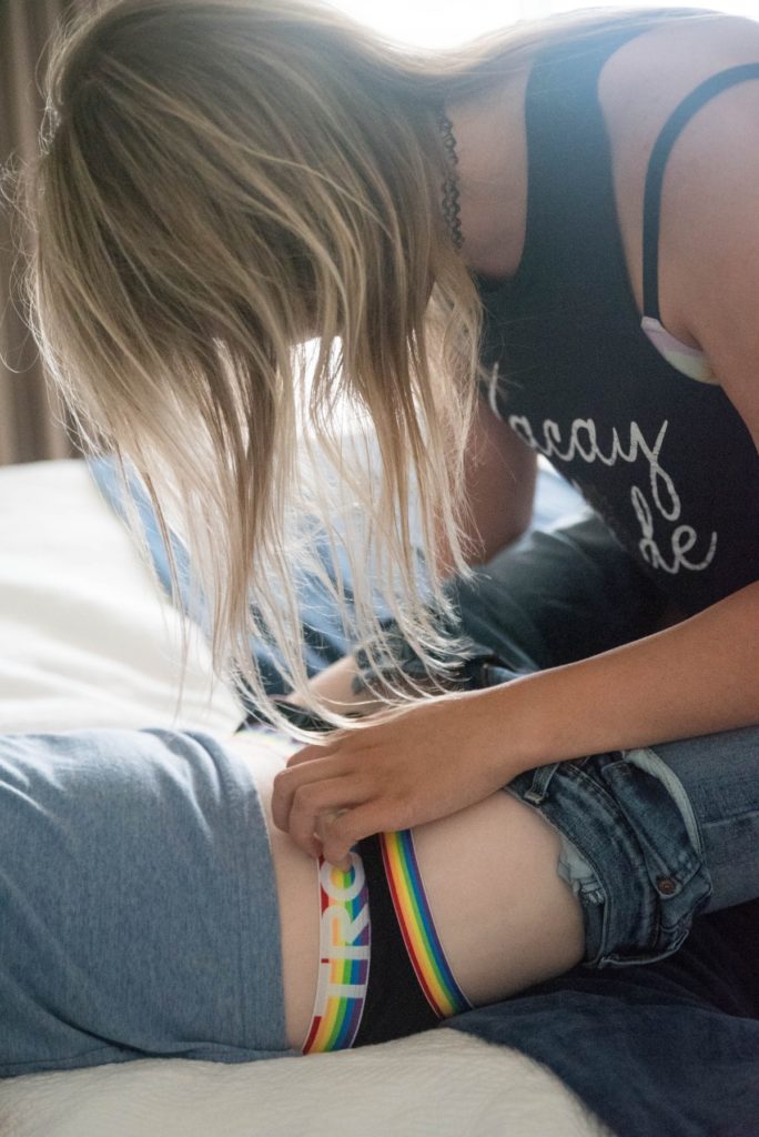 A person with long hair is kneeling between the knees of a guy in jeans who is laying down flat. They are unzipping the laying person's jeans, and the laying person's briefs can be seen. Image for How to Do Foreplay for Him article.