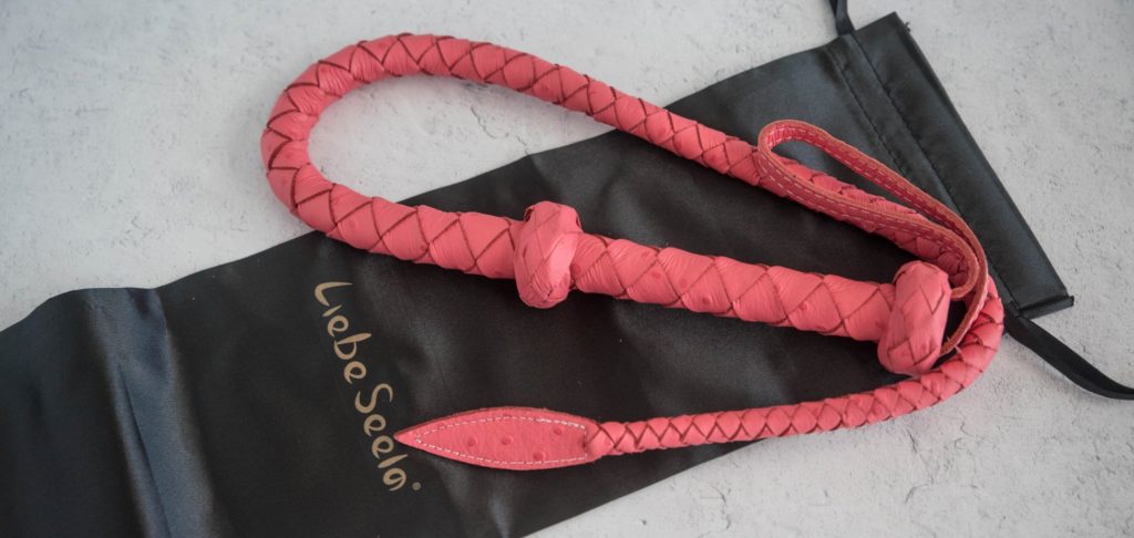 Top down view of the Angel's Kiss Whip. It's deep salmon color stands out against the black storage bag it is laying on. Image for Liebe Seele Angel's Kiss Whip review.