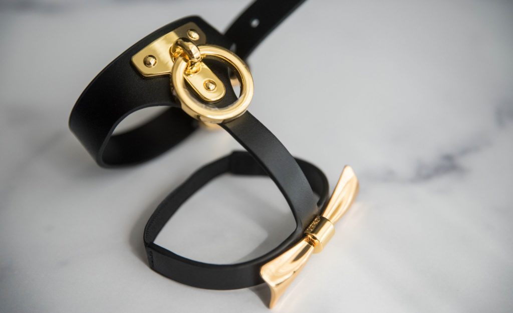One of the ankle cuffs sits out against a marbled white background. The black and gold contrast is stunning. This angle shows the entirety of the front of the cuff from the O-ring and its fastening plate to the metal gold bow on the opposite side. UPKO Butterfly Effect Ankle Cuffs review image.