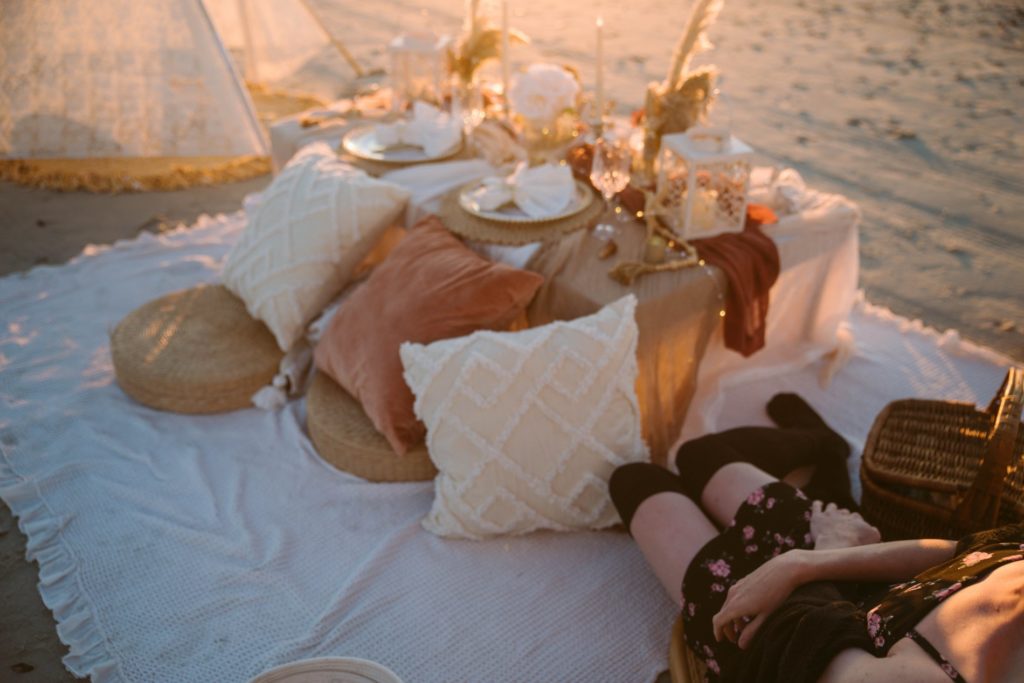 Image for my Sissy Date Ideas article. A slender person in a short dress is laying out in the corner of an image. The rest of the image shows an extravegant picnic set out on a beach while the sun is rising.