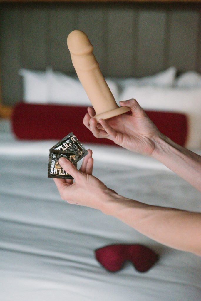 There is a hotel room bed with a red blindfold laying on the bed with condoms. Two hands in the forefront hold a realistic dildo and even more condoms. Image for my How to Fake a Threesome article.