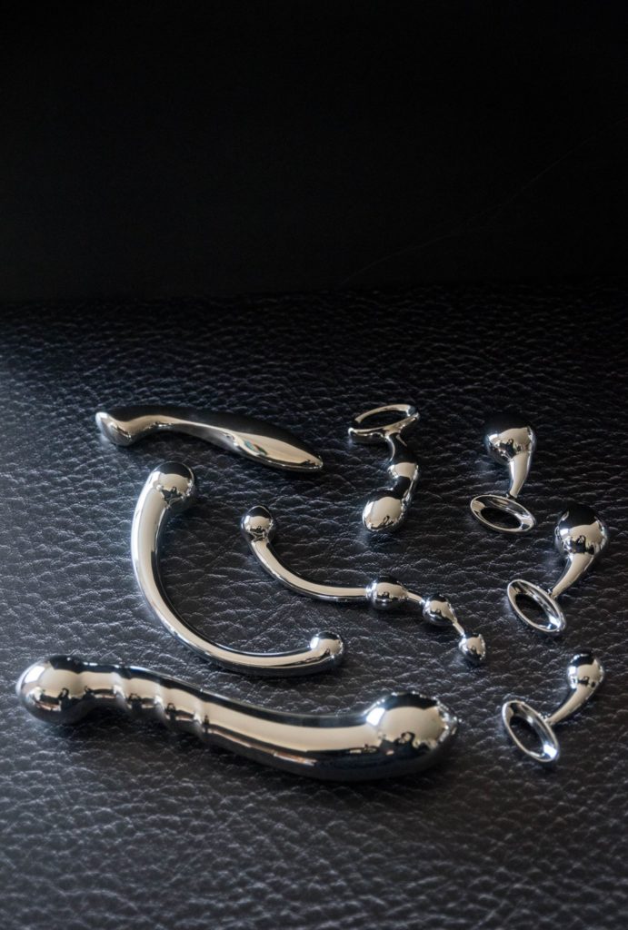 A spread of shiny, reflective stainless steel dildos laying out flat on black leather. The stainless steel sex toys almost glow in the dark in the shadowed lighting.