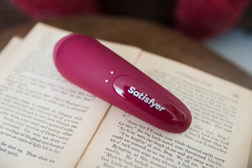 The Satisfyer Curvy 1 laying out on top of pages from an old book. The "Satisfyer" logo is very easily visible in addition to the magnetic charging ports for charging. This is a stylistic photo.