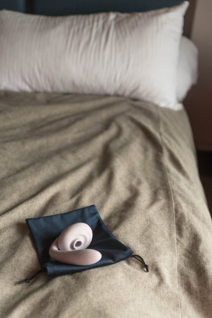 The Tracy's Dog OG Flow is resting on top of its included, blue drawstring bag on a bed. The bedding is a neutral light brown color. Natural light streams in from the right side of the room, bathing the vibrator in soft light. It looks welcoming. For my Tracy's Dog OG Flow review.