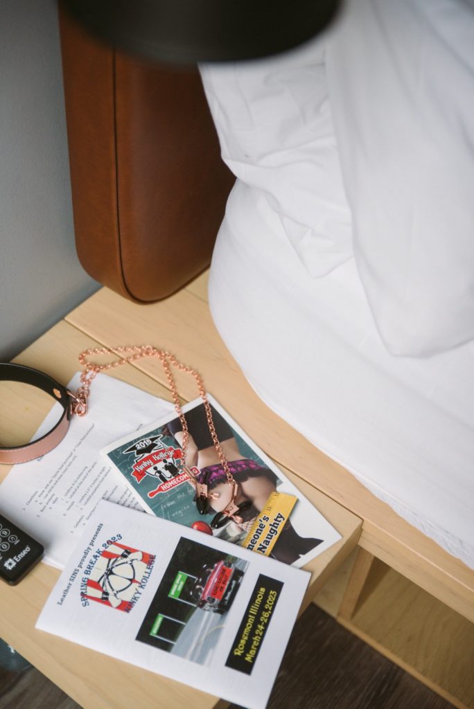 A collar, nipple clamps, a printed outline of class notes, and multiple BDSM convention booklets sit on a bedside hotel table. For my "BDSM Hotel Takeover: Everything You Need to Know" article.