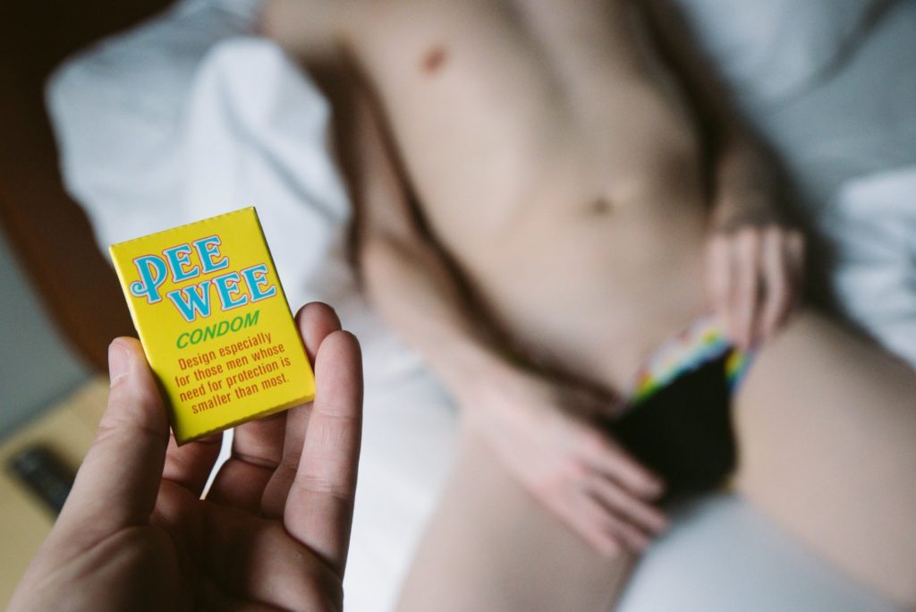 A person in their underwear lays on a bed. They are suggestively pulling down the waistband of their underwear, but nothing is showing. In the forefront, my hand holds a box that says "Pee Wee Condom. Design especially for those men whose need for protection is smaller than most." For my How to Give a Small Penis Humiliation Handjob article.