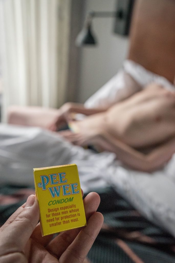 A person in their underwear lays on a bed. They are suggestively pulling down the waistband of their underwear, but nothing is showing. In the forefront, my hand holds a box that says "Pee Wee Condom. Design especially for those men whose need for protection is smaller than most." For my How to Give a Small Penis Humiliation Handjob article.
