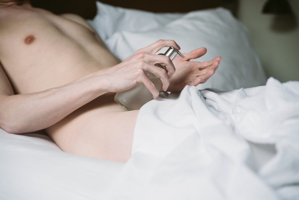 A person is laying naked on a bed. The white bedsheets are strategically placed to ensure nothing explicit is shown. They are holding a lube bottle in their hand, about to squirt lube onto their other, flattened hand. For my "how to find the best edging lube for hours of masturbation" article.