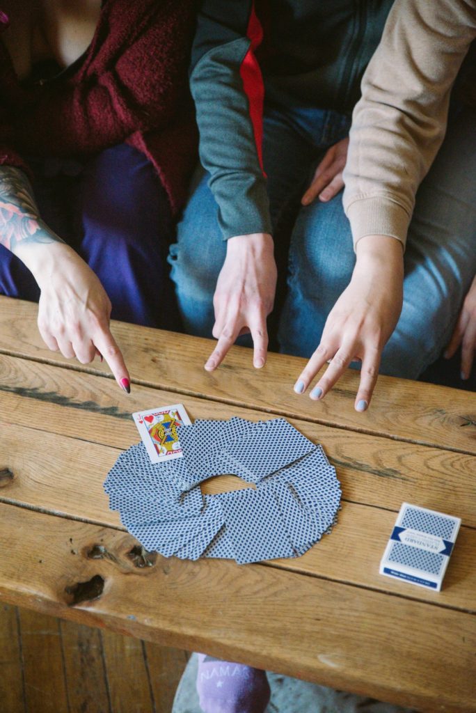 Three people sit on a couch, surrounding a table with a deck of cards spread out on the table. They have their fingers held up for the traditional Never Have I Ever counting game method. Image for my Femdom Never Have I Ever Questions article.