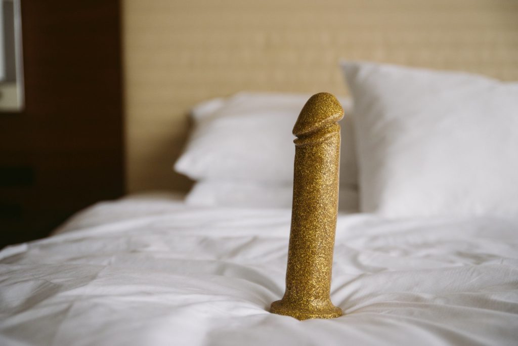 A golden dildo sits upright on white bedding. The headboard is in the background.Image for my How to Get a Huge Dildo into a Harness article.