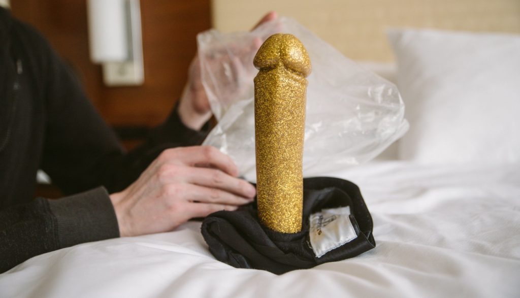 A golden dildo sits upright on white bedding. At the base is a strap-on harness folded up with the dildo protruding from the harness's built-in O-ring. A person holds a wrinkly plastic baggie in the background, right behind the dildo. The dildo looks large. Image for my How to Get a Huge Dildo into a Harness article.