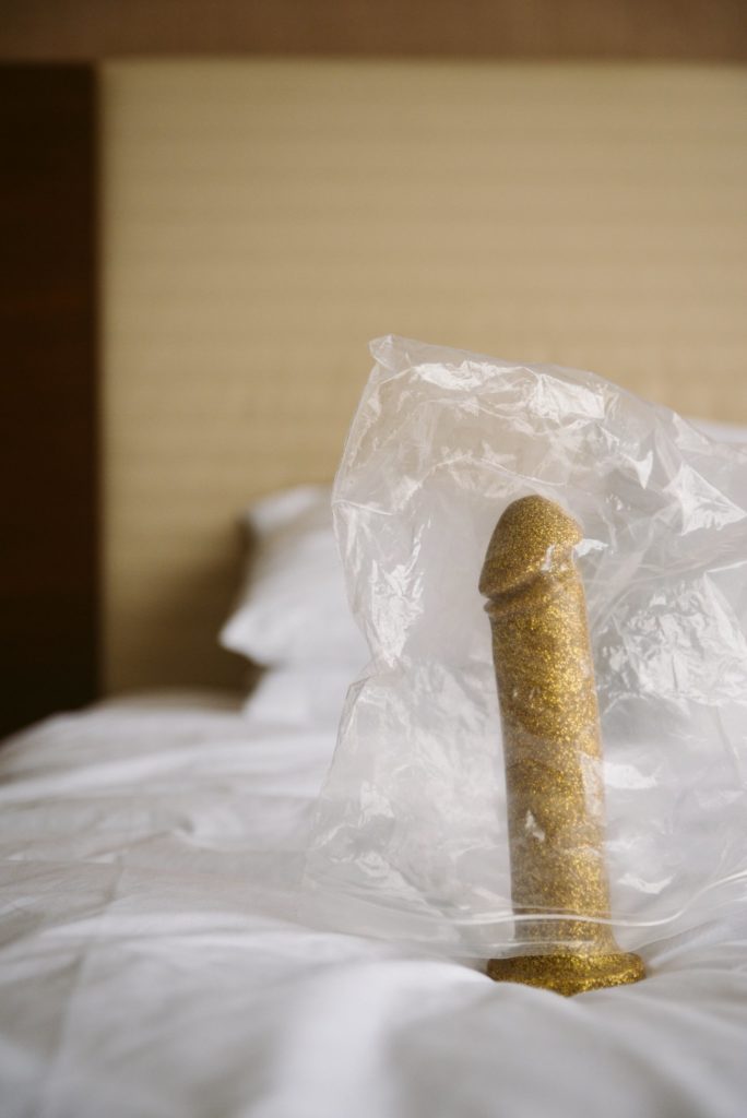 A golden dildo sits upright on white bedding. The headboard is in the background. A wrinkled plastic bag is loosely placed on top of the dildo, covering it like a weird blanket. Image for my How to Get a Huge Dildo into a Harness article.