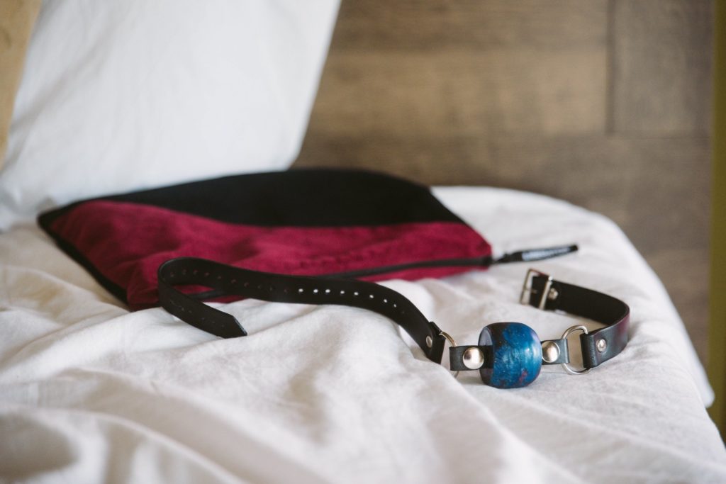 A colorful ball gag sits out on the white bedding of a bed with the headboard and wall in the background. There's a sex toy storage bag directly behind the ball gag. For my How to Keep Your Sex Life Private in an Apartment article.