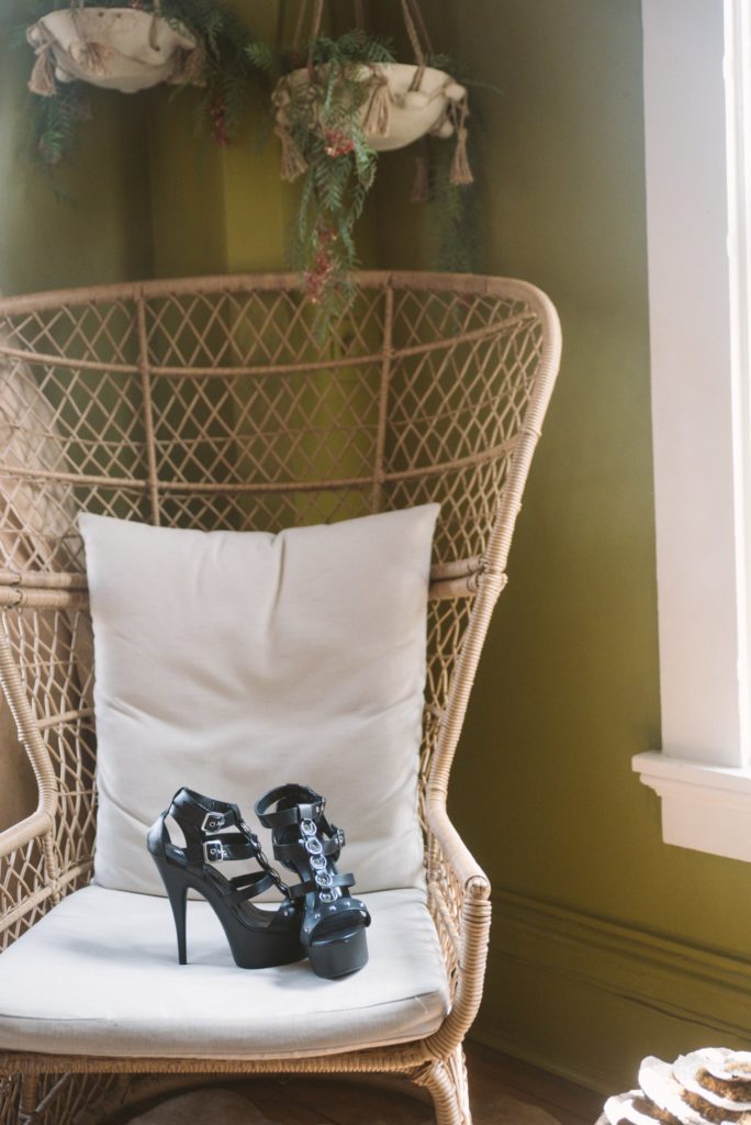 A pair of platform high heels sits on top of a wicker chair. The black heels stand out compared to the beige cushion. There are five O-rings that line the front of the heel. For my Lapdance Open Toed Studded Platform Heels review.
