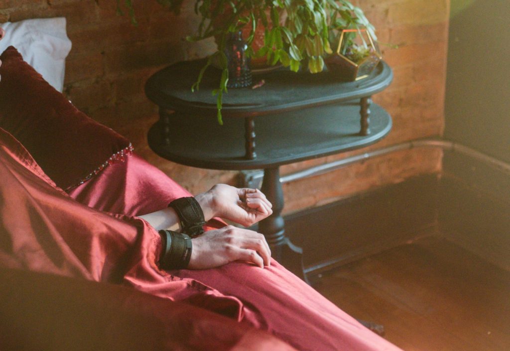 A person lays in a comfortable-looking bed with red bedding. They are mostly covered by the red blankets, but their hands stick out of the sides of one of the blankets. Their two hands are bound in nylon, velcro cuffs. There's a sunflare in the corner of the image since it's first thing in the morning. For my Sleeping in Bondage: How to Do It article.