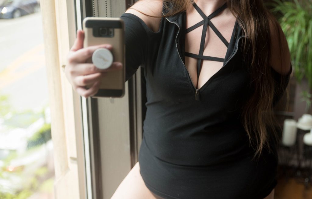 A person with long hair and a long shirt that shows off a lot of cleavage stands in front of a window. They're holding a cell phone in their hands with the front-facing camera pointed towards them. For my Tips for Taking the Perfect Sexting Photo article.