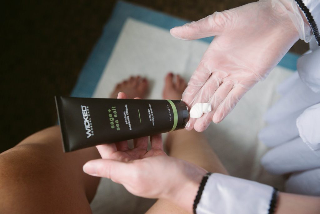 A person wearing a glove is squirting Wicked Sage and Sea Salt massage cream onto their gloved hand. Another person's tanned feet are shown in the background. The cream comes out as a cohesive cream - not an oil or anything runny. For my How to Give a Femdom Foot Massage article.
