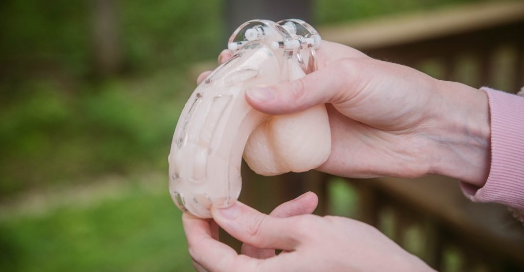 A person holds a flaccid packing dildo with one hand. With the other, they're sliding the shaft cage overtop of the shaft of the dildo, ensuring it meets the base ring at the bottom. For my How to Put on a Chastity Cage article.