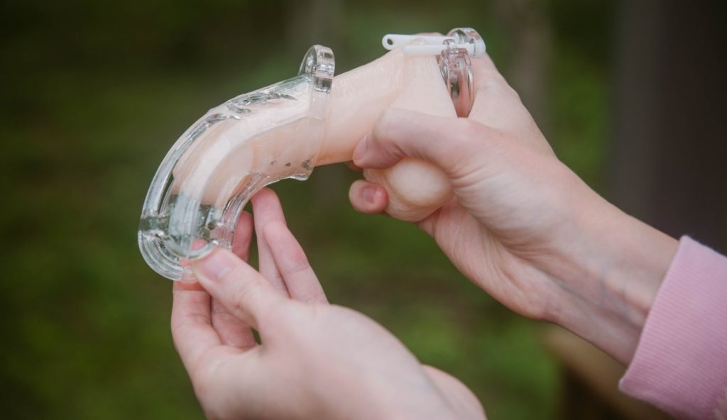 A person holds a flaccid packing dildo with one hand. With the other, they're sliding the shaft cage overtop of the shaft of the dildo, slowly pushing it towards the base ring. For my How to Put on a Chastity Cage article.