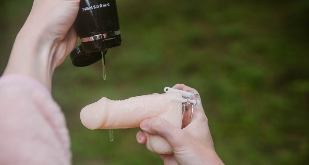 A person holds a flaccid packing dildo with the base ring of a chastity cage underneath the "balls" of the toy. They use their other hand to drizzle water-based lube onto the toy. For my How to Put on a Chastity Cage article.