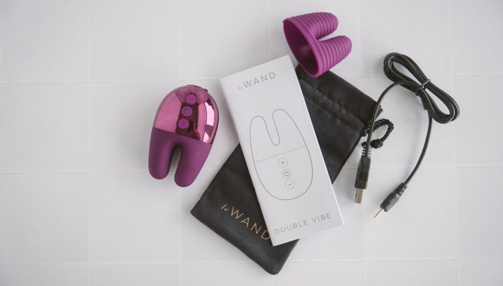 Everything included with the Double Vibe vibrator is laying out on a countertop. There's the vibrator, a textured cap, instruction manual, the charging cable, and the drawstring bag. For my Le Wand Double Vibe review.