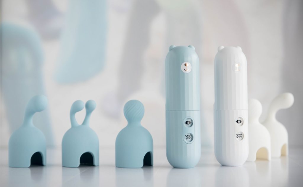 The blue and white MerBear vibrators are sitting upright on a surface with all of their attachment tips laid out next to them. This showcases the two different colors of the two toys when placed next to one another.