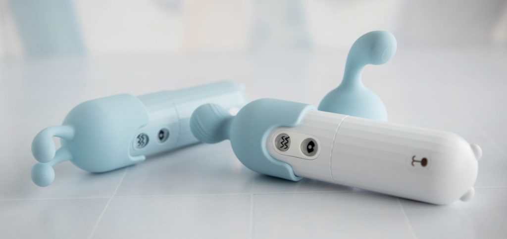 A MerBear vibrator in blue and white both laying sideways on a table. They both are "wearing" attachment tips, showcasing how the attachment tips have a cut out semi-circle pattern that allows all of the buttons to still be accessible while the attachment tips are on the toy.