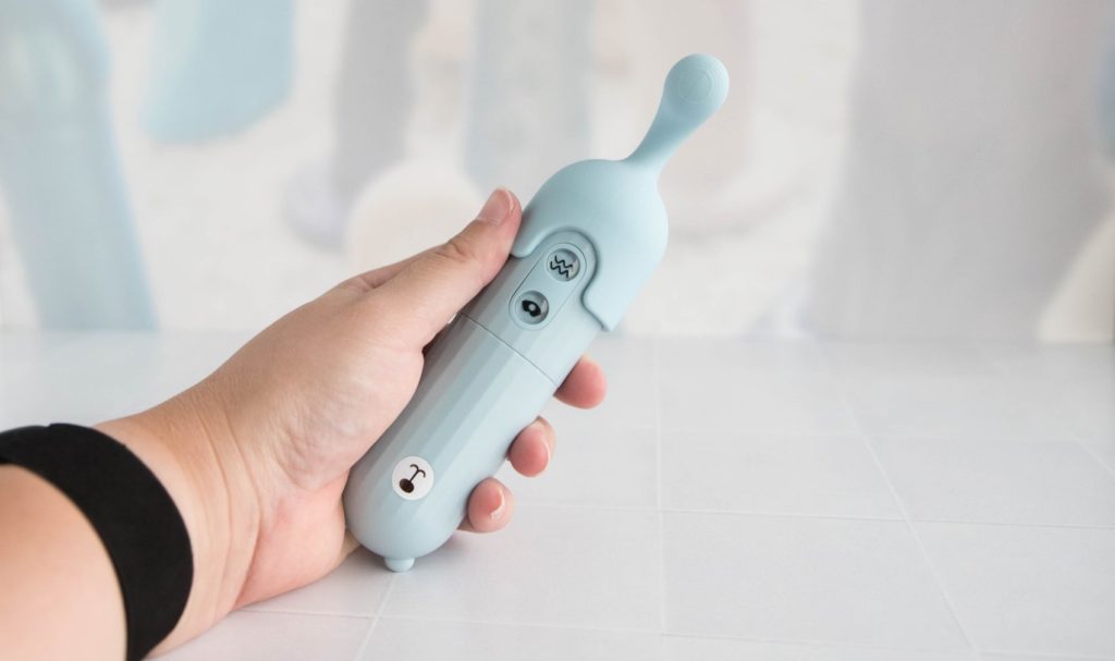 My hand holding the MerBear vibrator. It's slightly longer than my curled up hand around it. An attachment tip is shown on top of the vibrator, extending its length. My thumbs look like they could hit the control buttons with ease. For my MerBear review.