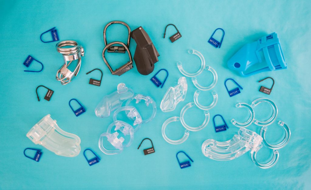 Multiple chastity cages and disposable plastic locks laid out on top of a blue background. Article for my Femdom Locktober Calendar.