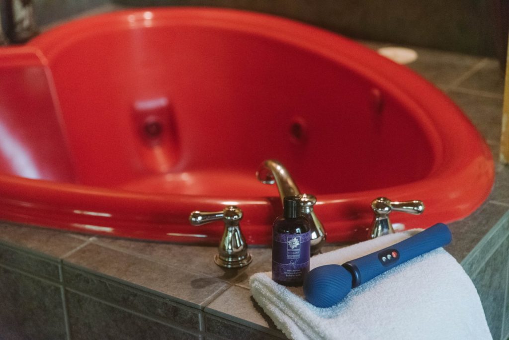 A wand massager and a bottle of vagina-friendly bubble bath are sitting on the edge of a tub on top of a white towel. The bathtub, shown in the background, is a large heart-shaped red bathtub. Image for my BDSM Scenes, Step-by-Step: Femdom Bath Slave article.