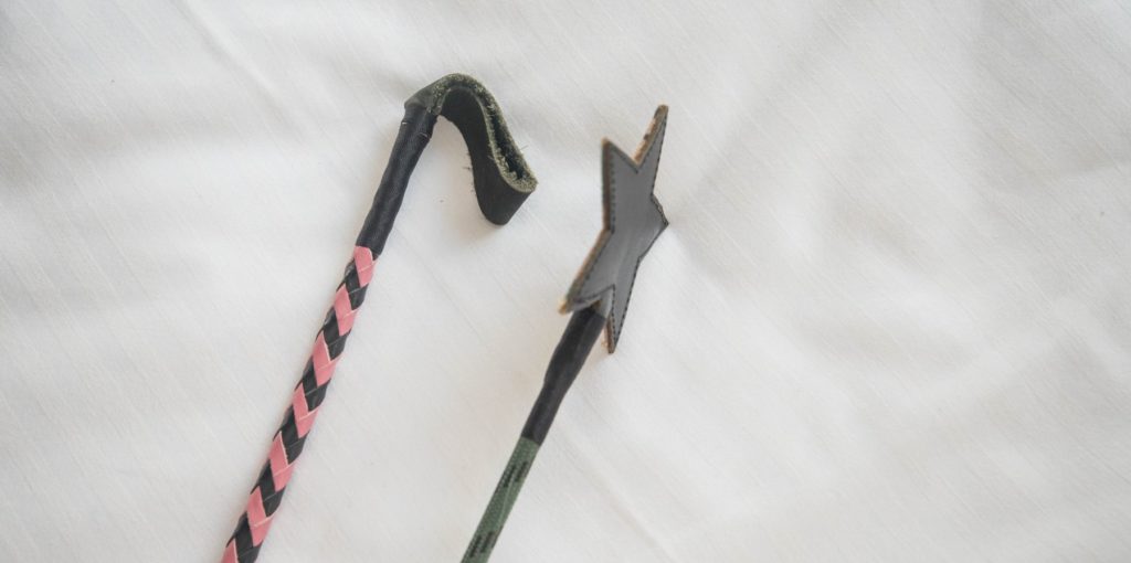 A close up of two tips of riding crops. The riding crop tip on the left is flexible and really floppy, folded in on itself. The riding crop on the right is completely rigid, holding its own shape up. For my riding crop for BDSM article.