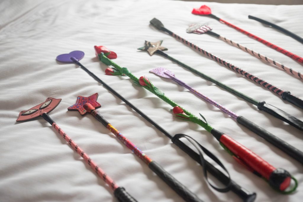 A whole collection of colorful riding crops laying out on top of a white bedspread next to an open window. For my riding crop for BDSM article.