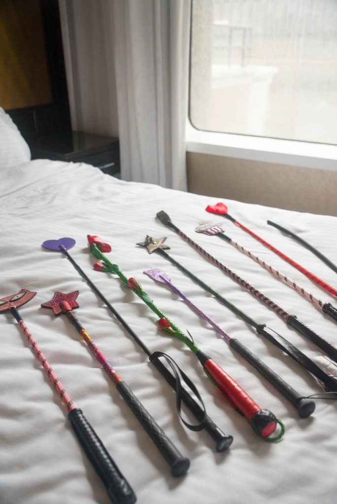 A whole collection of colorful riding crops laying out on top of a white bedspread next to an open window. For my riding crop for BDSM article.