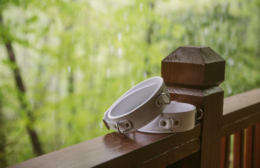 Phot of white thigh cuffs sitting out on a bannister of a wooden cabin. The green forest stands out beyond the balcony railing with rain pouring down from the trees behind the cuffs. For my Liebe Seele Fuji White Thigh Cuffs review.