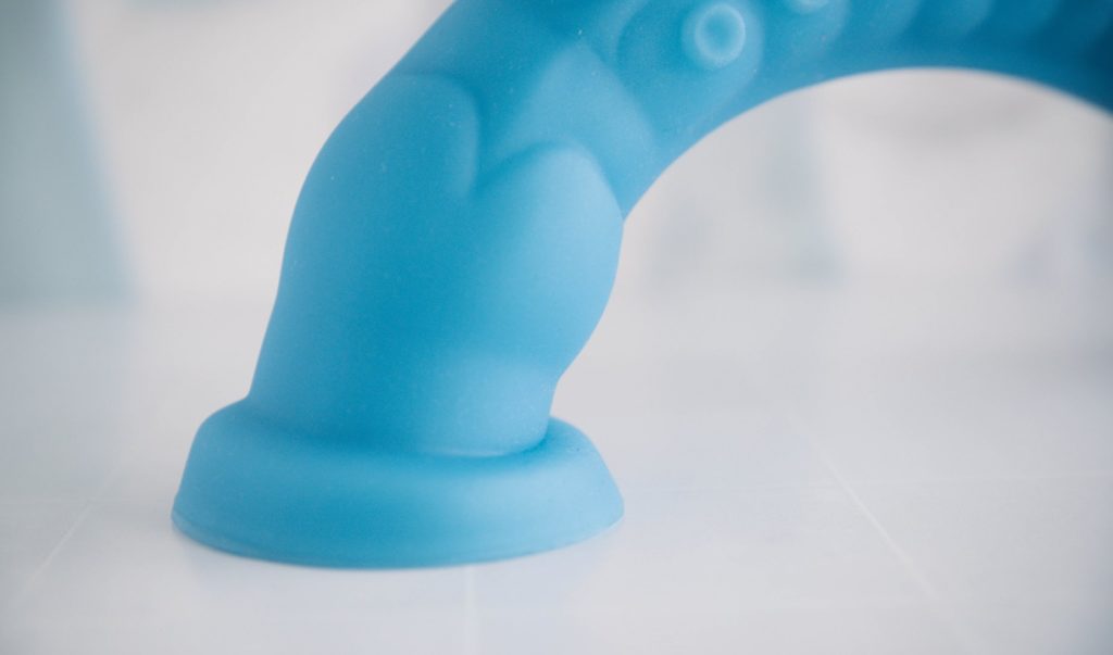 Close-up of the suction cup base on the bottom of the dildo. It is suctioned onto the floor underneath the dildo, and the close-up shows the bend in the suction cup that is helping it adhere to the surface beneath it.