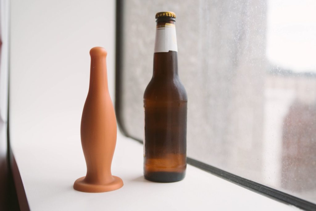 The beer bottle dildo sits on a windowsill, next to an unlabeled, standard beer bottle filled with beer. The Small size is noticeably smaller than the real-life beer bottle, but it has a similar shape. Photo for my MEO Dildo Bottle Butt Plug review.