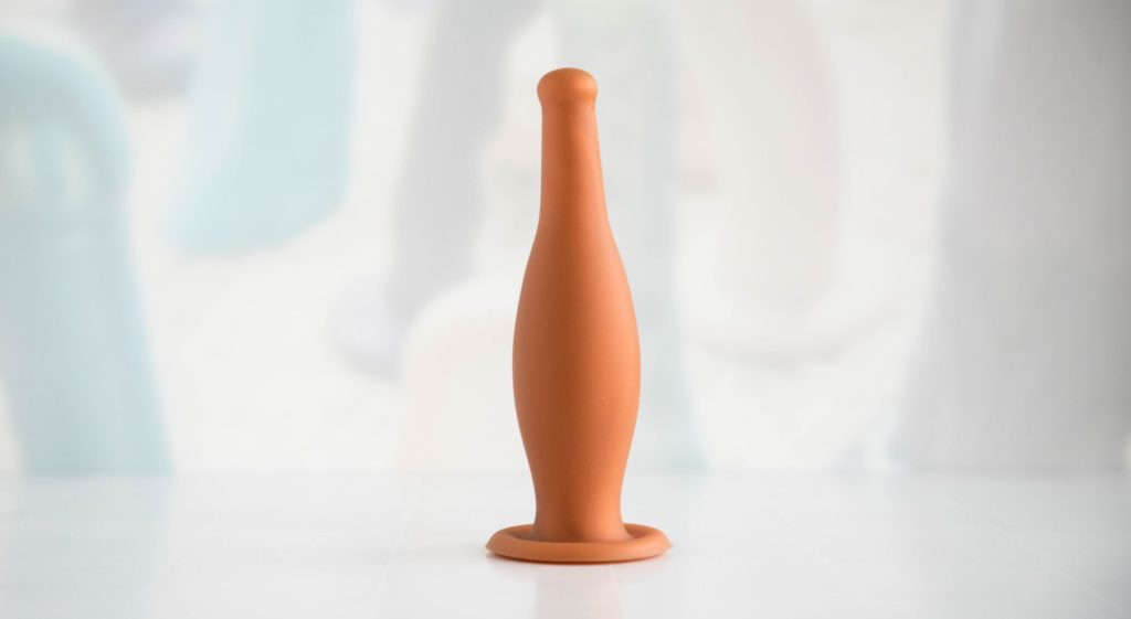 The MEO Dildo Bottle Butt Plug in front of a light background. It stands upright on its own. The suction cup base helps it with standing upright, and it's suctioned onto the table underneath it. It looks like an anal-safe version of a beer bottle.