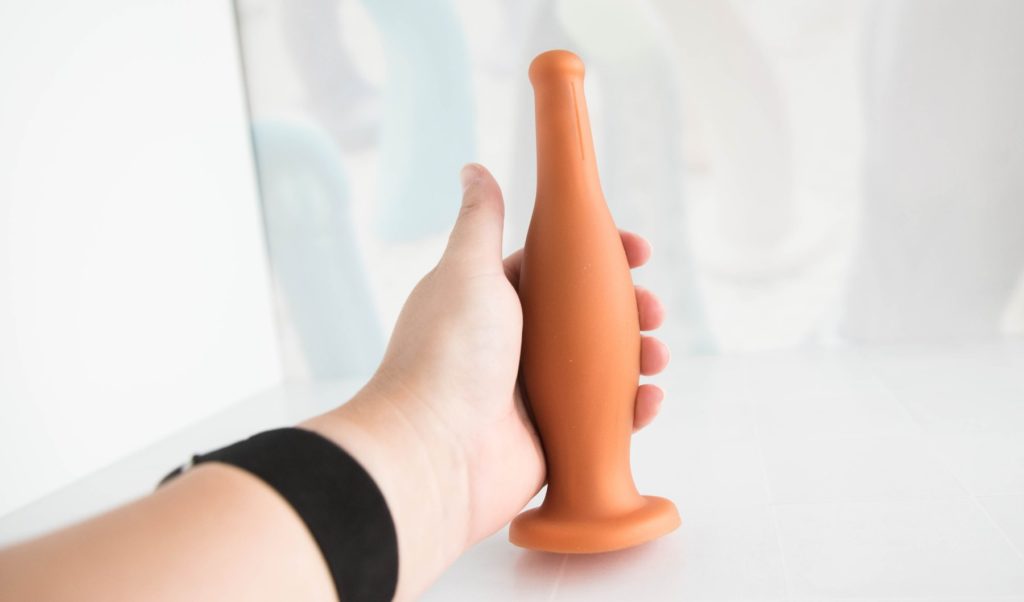 My hand wrapped around the Dildo Bottle Butt Plug in front of a light background. The toy almost looks like a genuine beer bottle in my hand, but it's slightly smaller. My fingers can't close around the "shaft" of the toy due to its thickness. Photo for my MEO Dildo Bottle Butt Plug review.