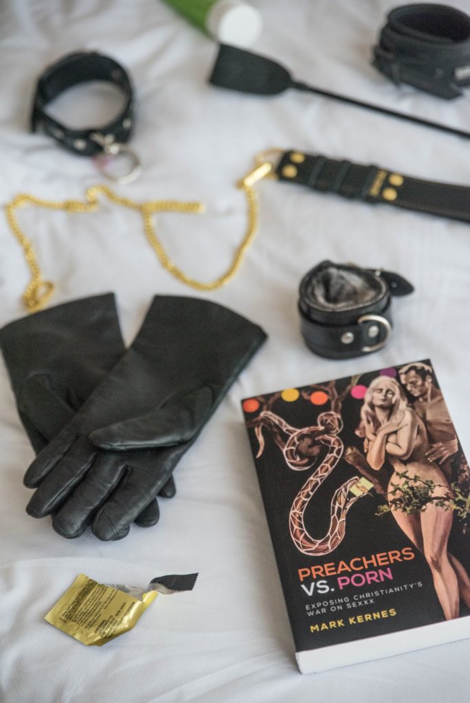 The book sits out on the white bedding of a bed during the daylight. It's surrounded by black and gold kinky sex things including gloves, cuffs, a leash, collar, riding crop, and empty gold condom wrapper. For my "Preachers Vs. Porn: Exposing Christianity's War on Sexxx" Review.