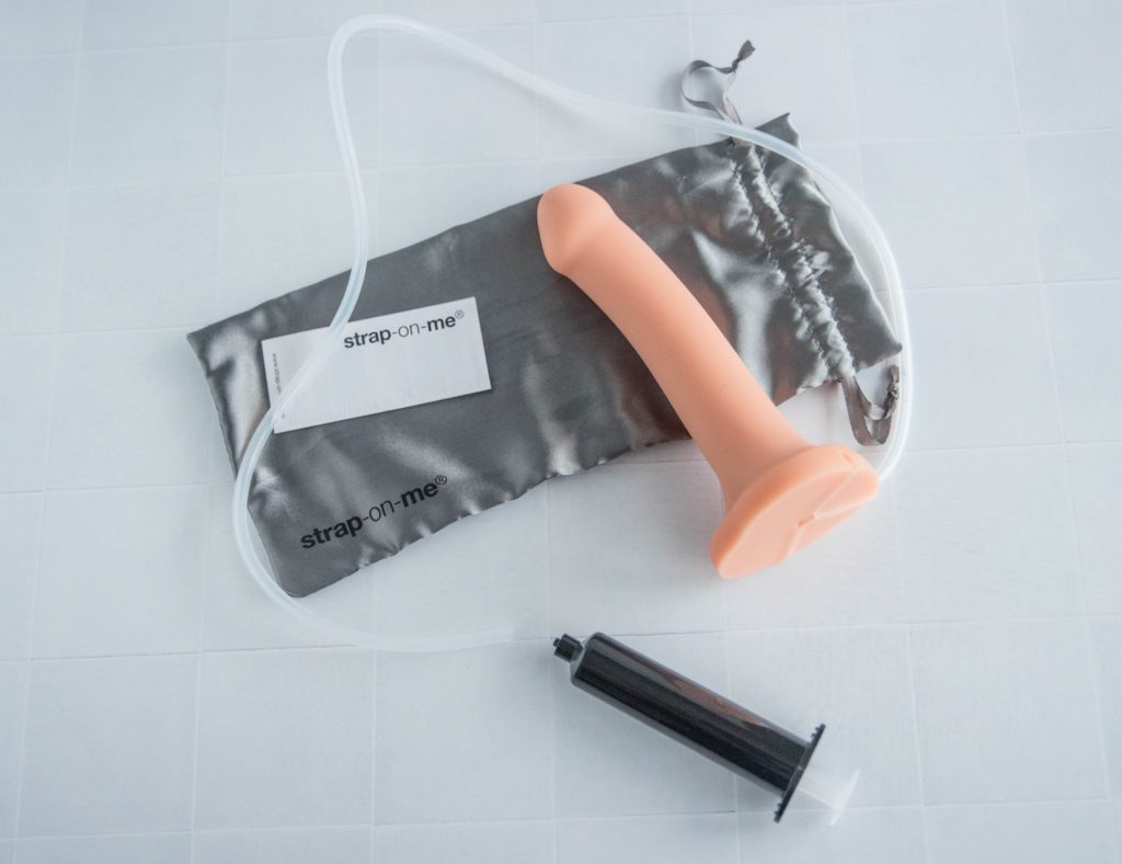 Everything included with the Silicone Cum Dildo laid out on a flat surface. It shows the dildo itself, the tubing next to the dildo, the syringe, the instruction manual, and the silver drawstring bag. The tubing is laid out around the entire image, showcasing how long the tubing is. Image for my Strap-on-Me Silicone Cum Dildo review.