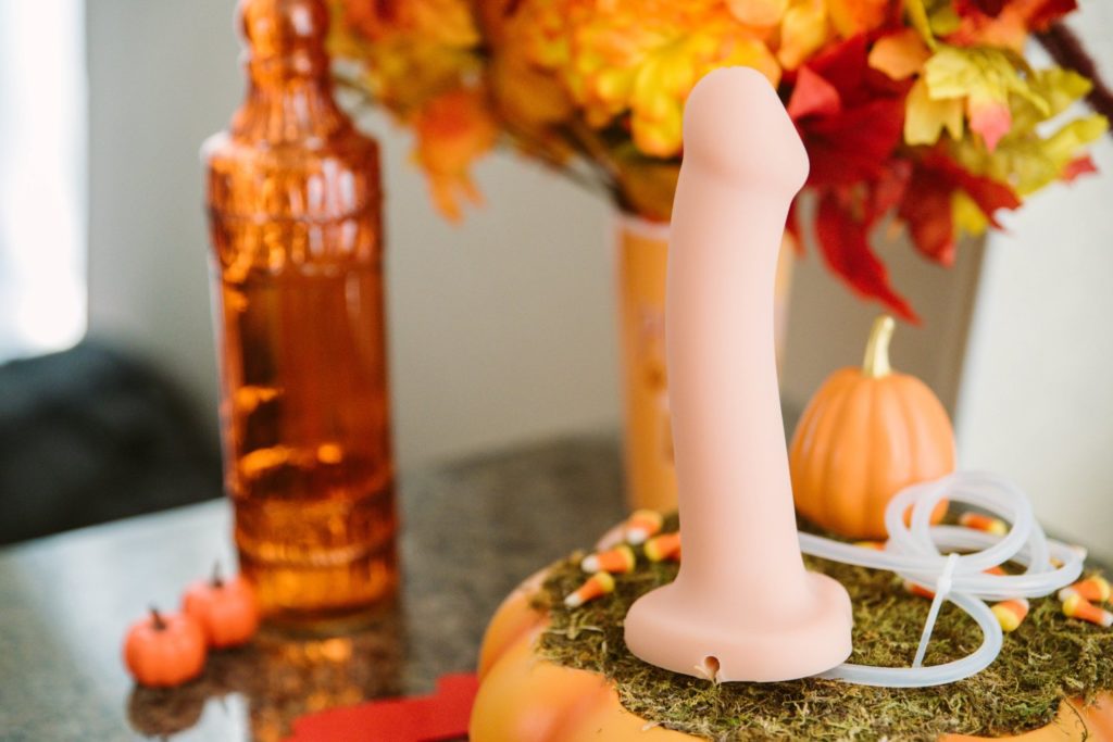 The Silicone Cum Dildo is sitting out on a grassy platform on top of a pumpkin on a kitchen countertop. It is surrounded by candy corn. In the background, there is red, yellow, and orange flowers and other orange and fall items. The photo looks very fall seasonal. For my Strap-on-Me Silicone Cum Dildo review.