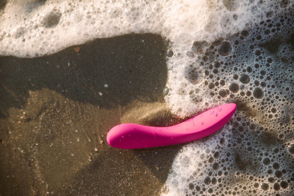 The We-Vibe Rave 2 sitting in the sand. A wave from the ocean has just rolled in, and the bubbles and ocean water making beautiful textures all around the vibrator. There are droplets of ocean water covering the vibrator. Image for my We-Vibe Rave 2 review.