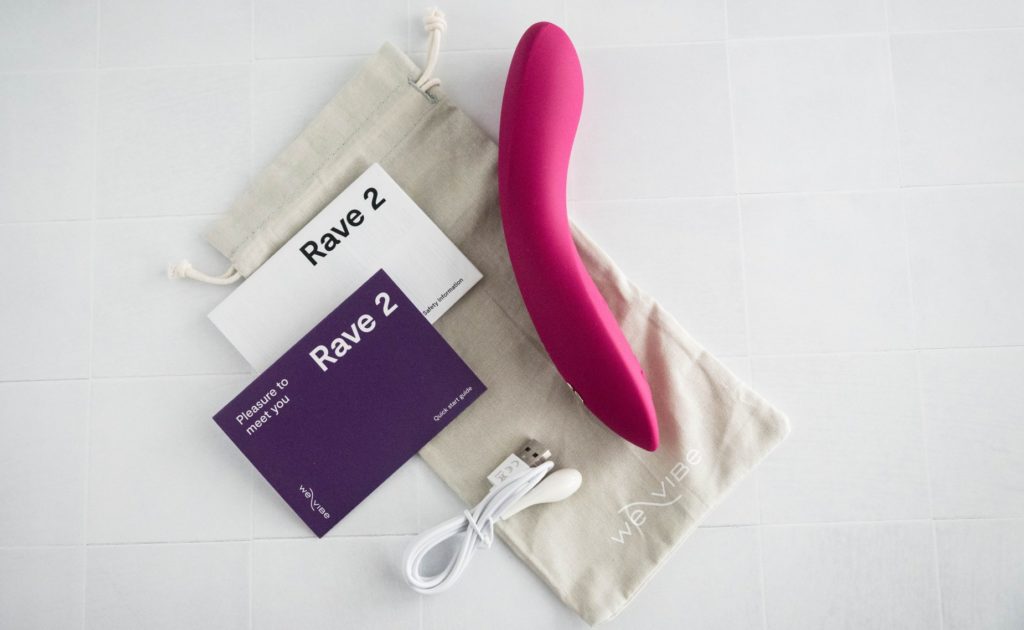 The We-Vibe Rave 2 and everything it comes with laying out flat in this top-down image. There's the pink vibrator itself, a quick start guide, the detailed instructions, the magnetic charging cable, and a canvas drawstring storage bag for the vibrator.