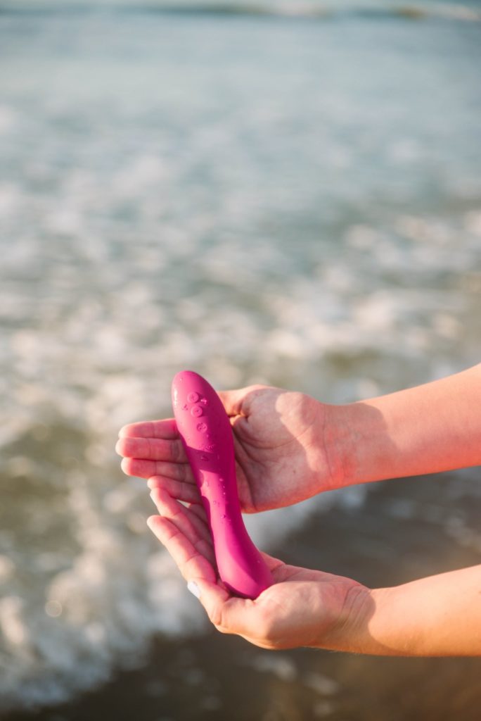 Two hands cradle the pink vibrator. In the background, there's a wave from the ocean coming in in the background, underneath the person's outstretched hands. Image for my We-Vibe Rave 2 review.
