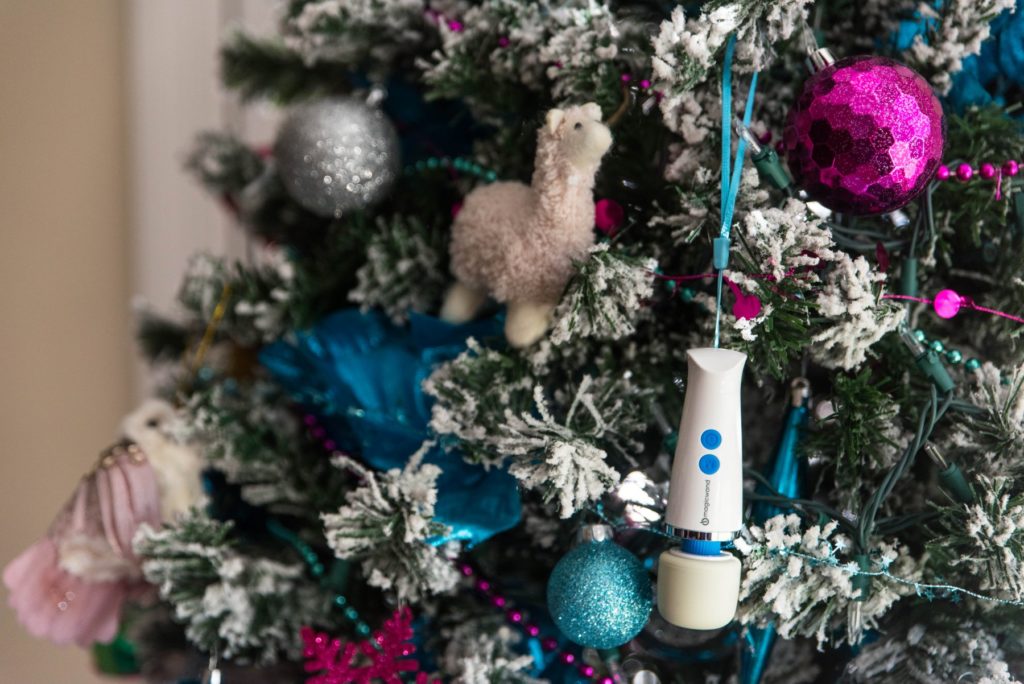 Small vibrator hanging by a blue wrist strap on a Christmas tree. The tree is flocked with pink and blue ornaments. The Magic Wand stands out against the tree's branches. Image for my Vibratex Magic Wand Micro Review.