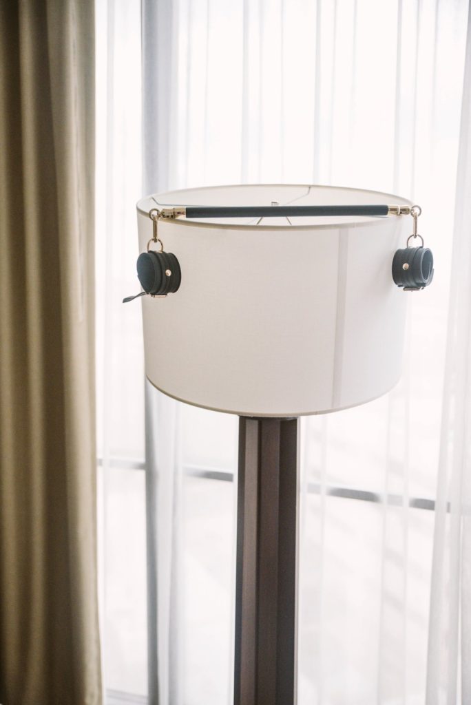 The spreader bar is resting on top of a large, floor lamp's lampshade. Its length almost perfectly matches the width of the lampshade. Two cuffs hang off the end of the spreader bar. Image for my Liebe Seele Black Organosilicon Spreader Bar Review.