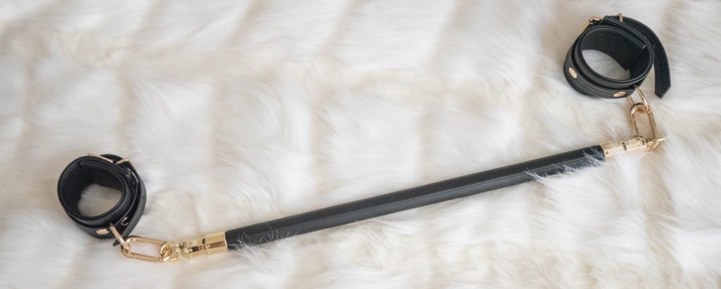 The spreader bar, including its two gold clips, laying out on top of a faux fur blanket. It looks luxurious and soft. Separately sold, color-matching Black Organosilicon wrist cuffs are attached onto the ends of the spreader bar. Image for my Liebe Seele Black Organosilicon Spreader Bar Review.