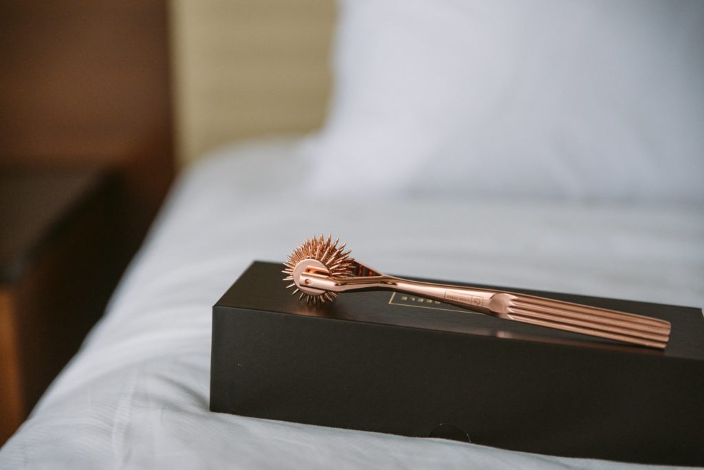 The rose gold pinwheel sits out on top of its black box on top of a bed with white bedding. The headboard is visible in the background, and the spikes of the pinwheel look extremely sharp. For my Liebe Seele X-Rose Wartenburg Pinwheel review.